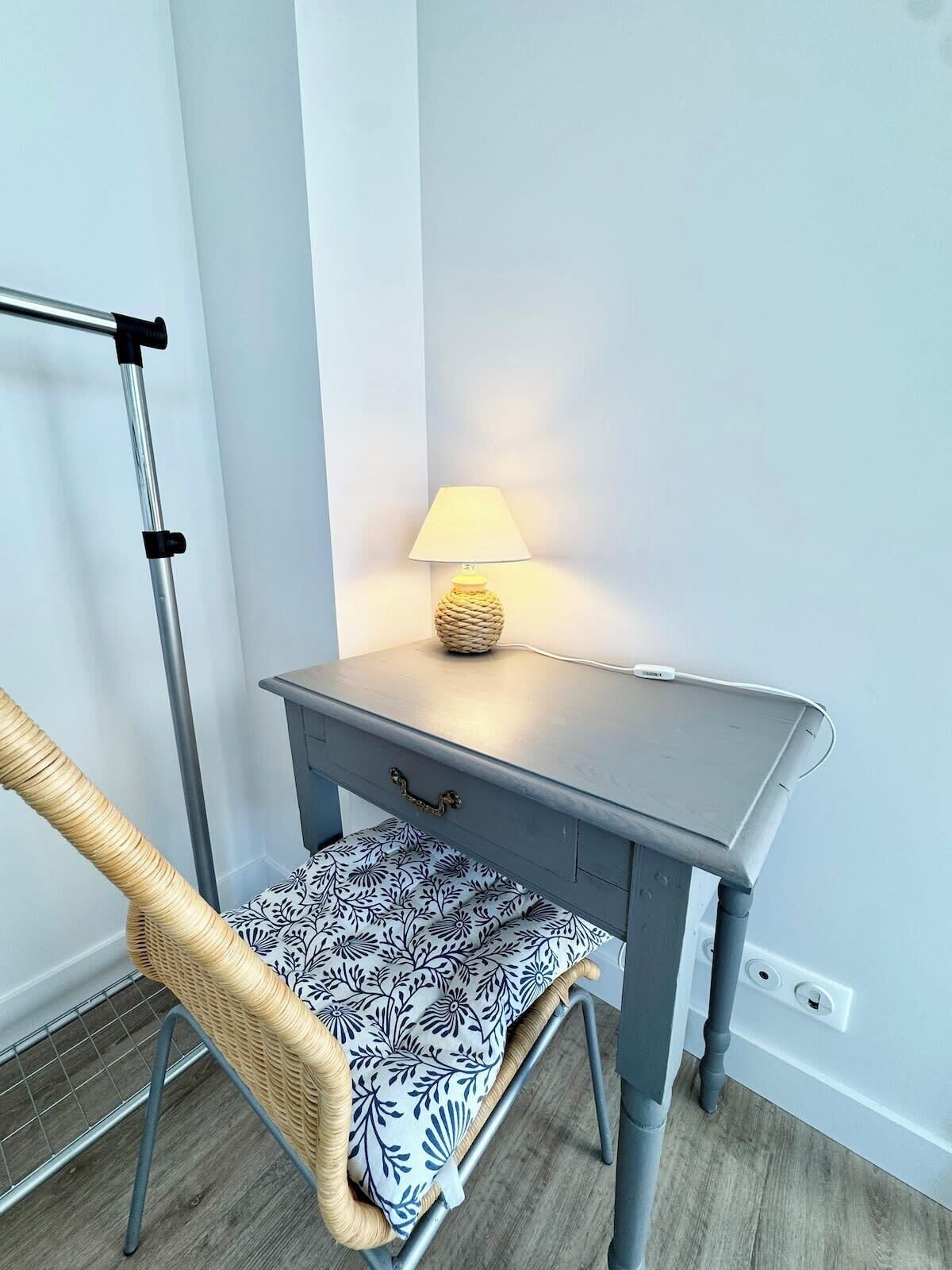 Ett litet, grått skrivbord med en lampa, en flätad stol med ett mönstrat kuddöverdrag och ett metallstativ.
