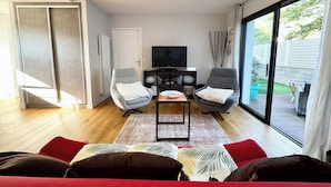 A modern living room with a flat-screen TV, two grey armchairs, a red sofa, and a glass door leading to an outdoor area.