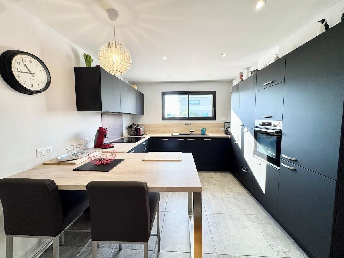 Una cocina moderna con comedor, que incluye una mesa de madera, sillas negras, un reloj negro y una lámpara colgante.