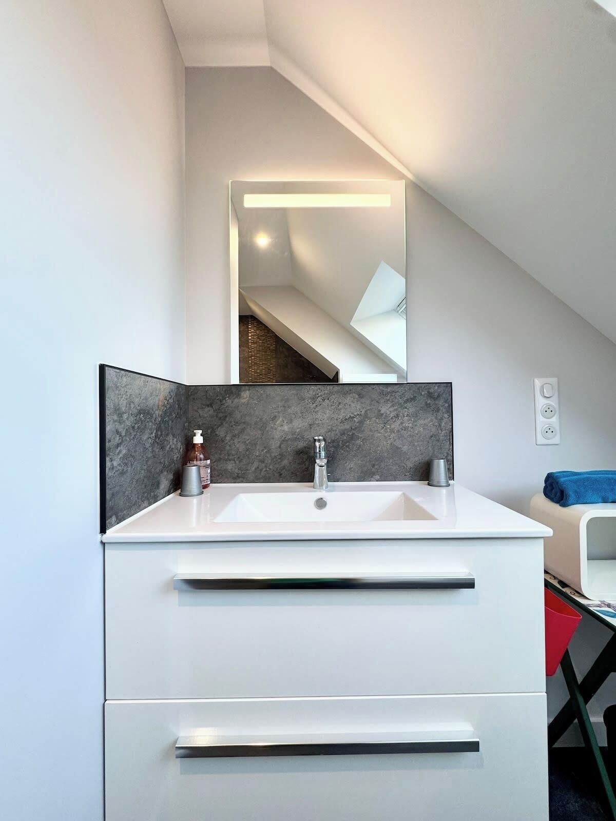 Un baño moderno con tocador blanco, un espejo grande y un protector contra salpicaduras de mármol oscuro.