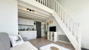 A modern living room with a sofa, a coffee table, a staircase, a kitchen, and a flat-screen TV. - Superbe appartement duplex - Vue mer - Piscine (Guidel)