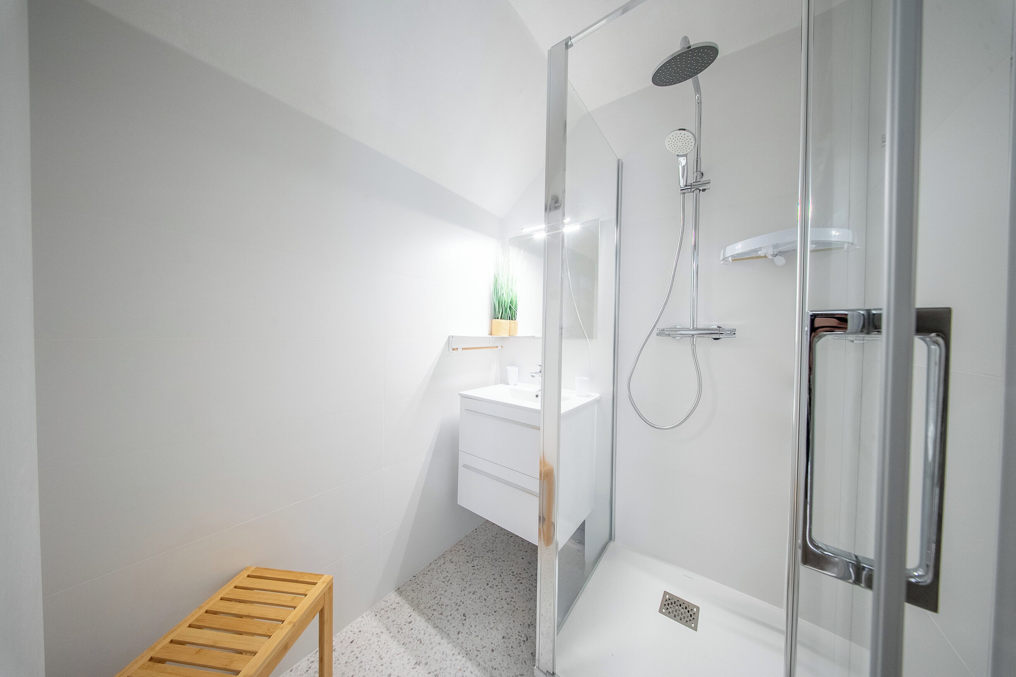 Une salle de bain moderne avec une douche à parois de verre, un banc en bois et un lavabo blanc agrémenté d’une plante.