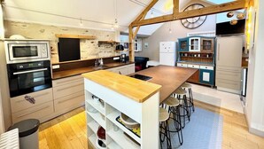 A modern kitchen with wooden beams, a central island, bar stools, and built-in appliances. - Spacieuse longère archi cosy - Jardin (Quimperlé)