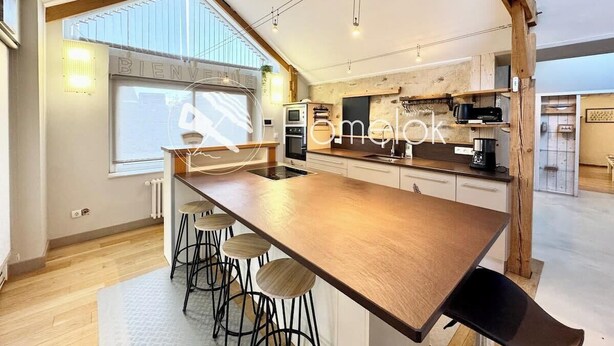 A modern kitchen with a central island, bar stools, and a large window providing natural light. - Spacieuse longère archi cosy - Jardin (Quimperlé)