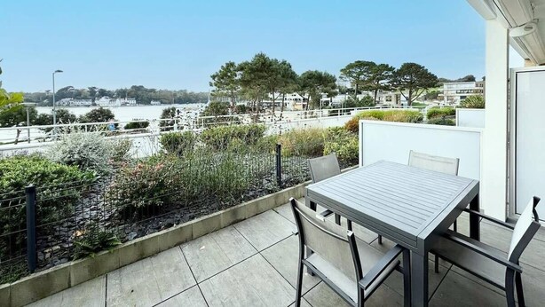 Balcón con mesa y sillas que da a la orilla con vista a edificios y vegetación.