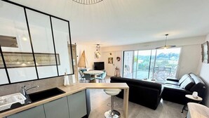 Una cocina moderna con una isla integrada y taburetes de bar, una zona de comedor con una mesa redonda y una sala de estar con sofá y vistas al exterior.