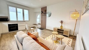 A modern living room with a sofa, a small table, and a lamp. There is a kitchen area with a stove and cabinets. - Lovely ! T2 neuf archi cosy 1 (Quimperlé)