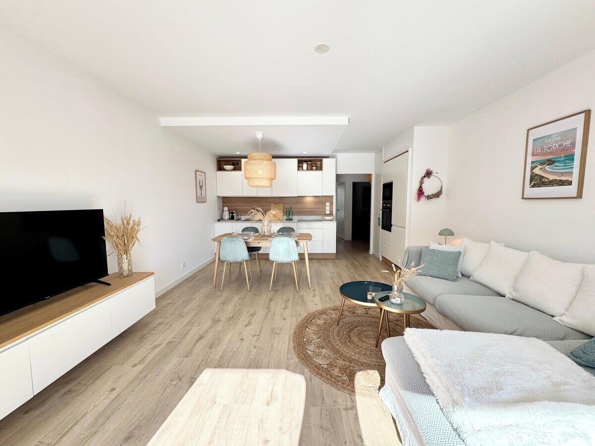 Ein modernes Wohnzimmer mit Fernseher, einer Couch, einem Couchtisch und einem Essbereich mit Stühlen, im Hintergrund befindet sich eine Küche.
