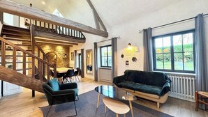 A spacious living area with a wooden staircase, a black sofa, a glass coffee table, and a view of the outdoors through large windows.