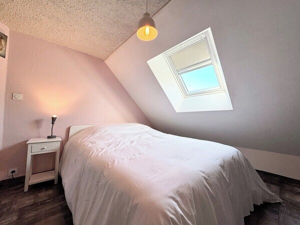 Un dormitorio con techo inclinado, una cama, una mesita de noche, una lámpara y una ventana con vistas.