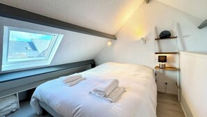 Une chambre d’hôtel avec un lit simple, une lucarne, une table de chevet avec une lampe et une étagère fixée au mur.