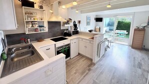 A modern kitchen with white cabinetry, a built-in oven, and a large window overlooking a garden. - Maisonnette 300m des plages - Piscine chaufée (Guidel)