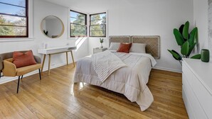 A modern bedroom with a large bed, a desk, and a chair. There are windows providing natural light and a view of trees.