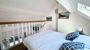 Un dormitorio con techo inclinado, una cama con ropa de cama blanca, una mesita de noche con lámpara y una ventana con vista a un paisaje.
