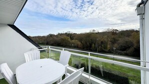 A balcony with a white table and chairs, offering a view of a road and trees.