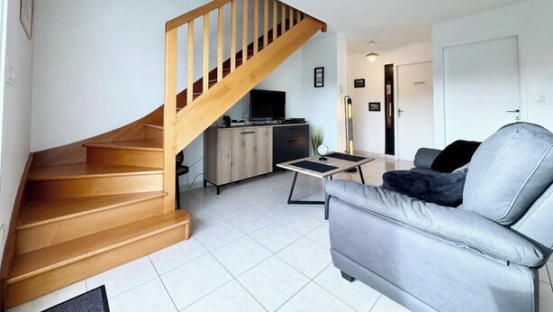A modern living room with a wooden staircase, a grey sofa, and a small kitchen area.