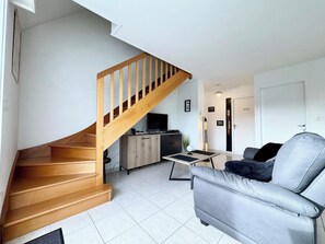 A modern living room with a wooden staircase, a grey sofa, and a small kitchen area.