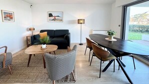 Un salon moderne avec un canapé noir, une table basse ronde en bois et une table à manger avec des chaises. Des tableaux encadrés ornent le mur, ainsi qu’une lampe sur pied. Une grande fenêtre offre une vue sur l’extérieur.