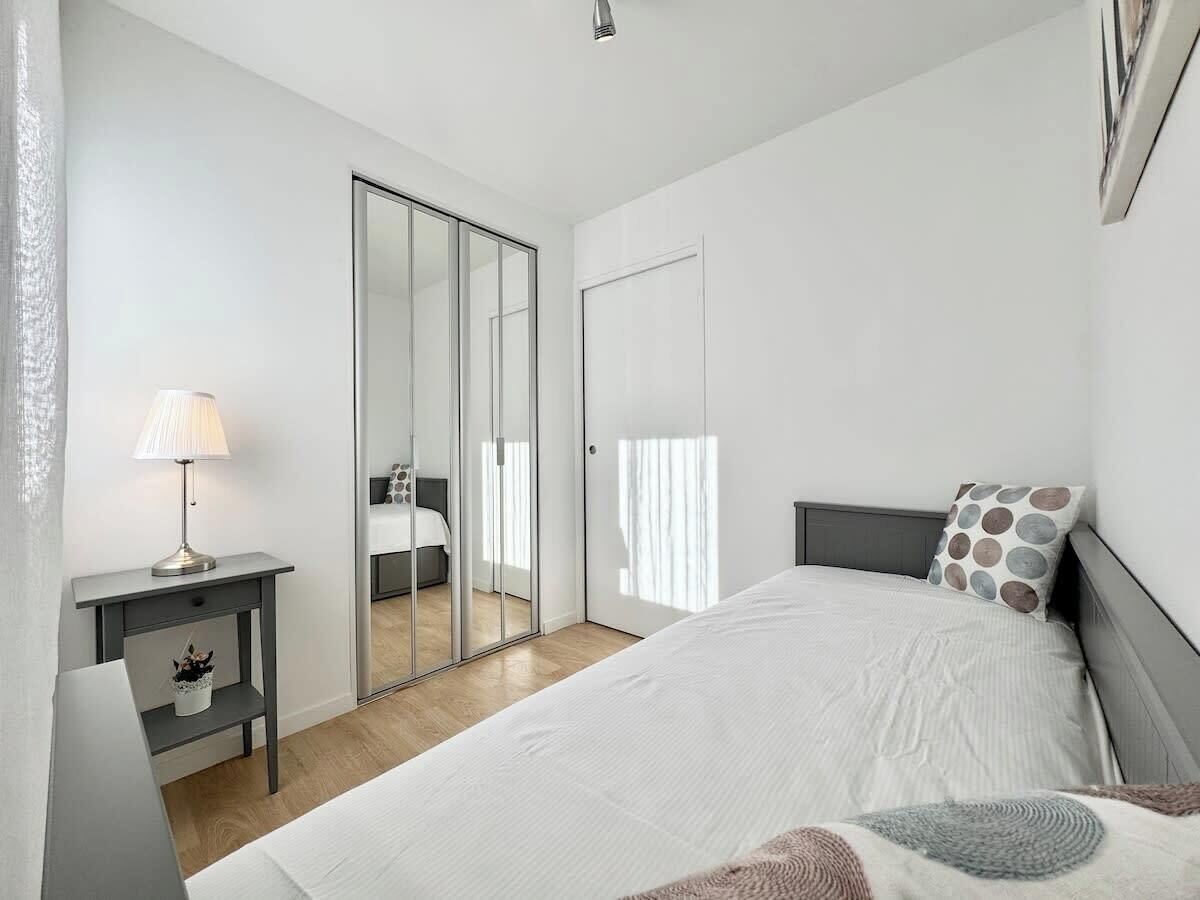 Un dormitorio con cama, mesita de noche, lámpara, espejo y puerta corredera que da a otra habitación.