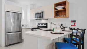 A modern kitchen with a stainless steel refrigerator, microwave, and oven. There is a wooden shelf above the sink holding glasses and a bowl. A chair with a blue cushion is visible. - Spacious 2BR Condo w/ Free Parking near Palm Beach (Noord)