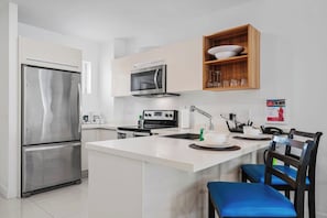 A modern kitchen with a stainless steel refrigerator, microwave, and oven. There is a wooden shelf above the sink holding glasses and a bowl. A chair with a blue cushion is visible. - Spacious 2BR Condo w/ Free Parking near Palm Beach (Noord)