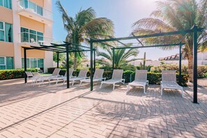 A covered outdoor seating area with white lounge chairs, palm trees, and a building with balconies.
