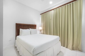 A hotel room with a bed, curtains, a nightstand, and a lamp. - Spacious 2BR Condo with Pool Access in Noord Aruba (Noord)