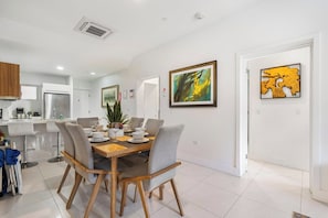 A modern dining area with a wooden table and grey chairs, a kitchen in the background, and two framed artworks on the walls. - Ground-Floor 2BR Condo w/ Patio & Pool, Noord (Noord)
