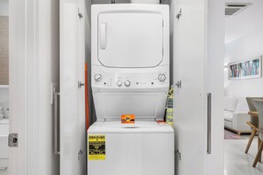 A compact, white washing machine with a control panel, placed in a modern, minimalistic room with a white door and a framed picture on the wall.