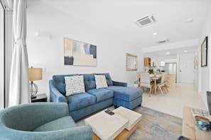 A modern living room with a blue sofa, a coffee table, and a dining area in the background. - Ground-Floor 2BR Condo w/ Patio & Pool, Noord (Noord)