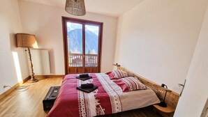 A bedroom with a wooden floor, a bed with a red and white patterned bedspread, a wooden headboard, a bedside table with a lamp, and a view of mountains through a window. - Guy and Rose's cottage (Passy)