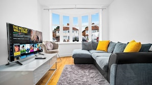 Una sala de estar moderna con un televisor de pantalla plana grande, un sofá gris, un sillón negro y vistas de edificios residenciales a través de un ventanal.