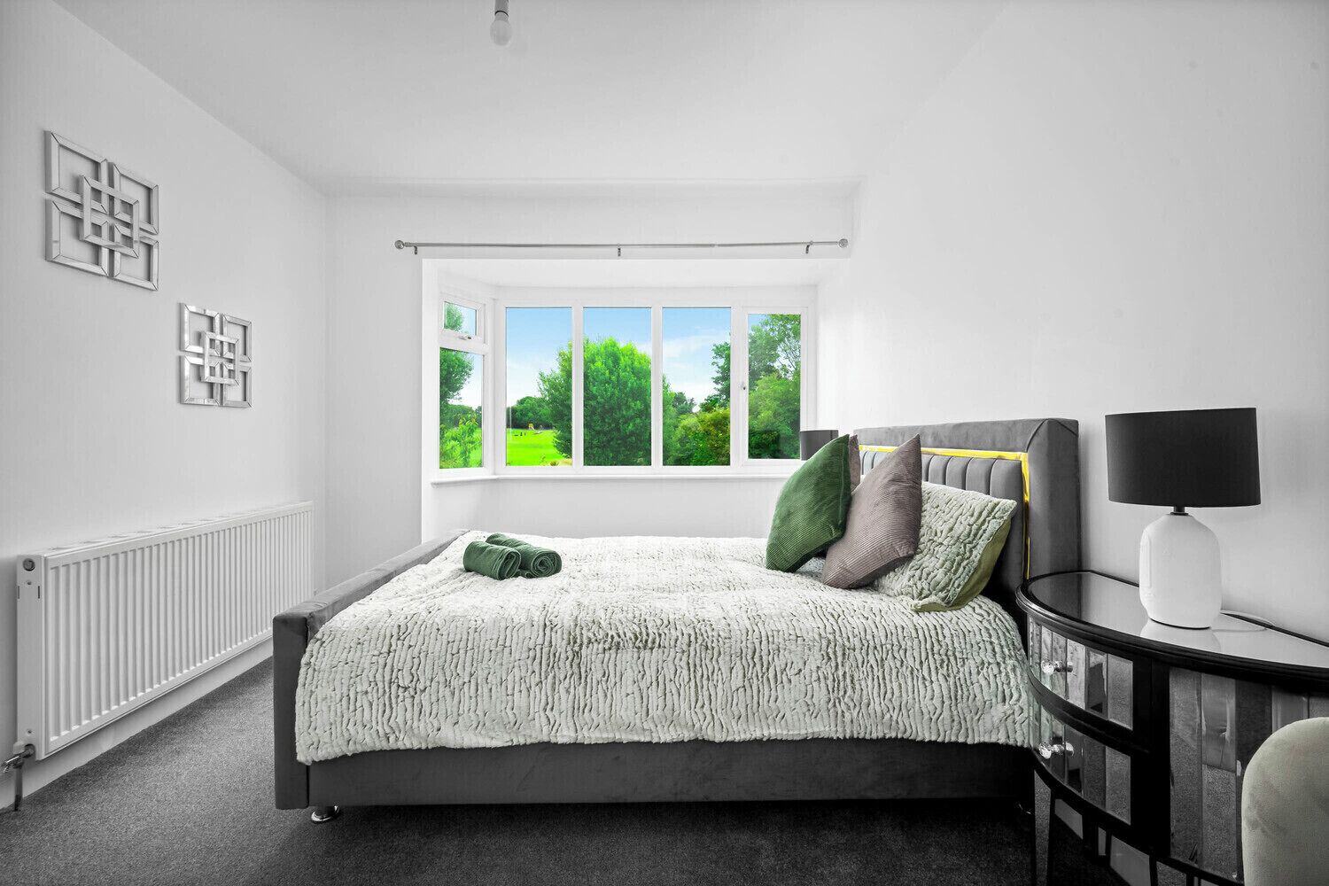 Et soveværelse med et stort vindue, en seng med gråt sengegavl, et natbord med en lampe og en radiator.