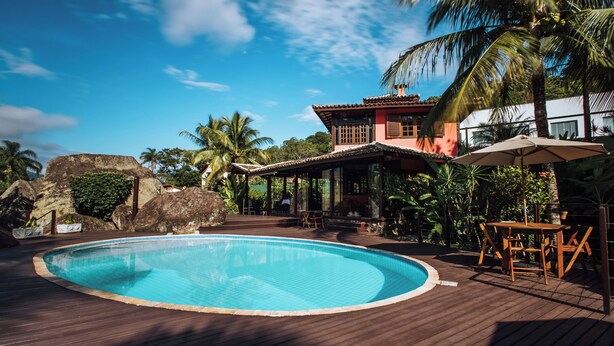 A swimming pool surrounded by a wooden deck, palm trees, and a building with a red roof. - Pousada Vila das Pedras (Ilhabela)