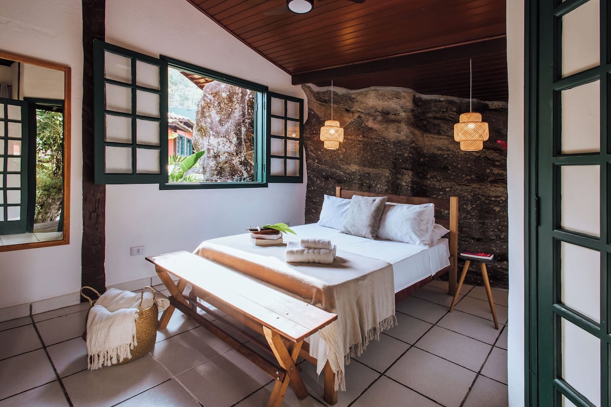 Un dormitorio con cama, un banco de madera, una cesta de mimbre y una pared de piedra.