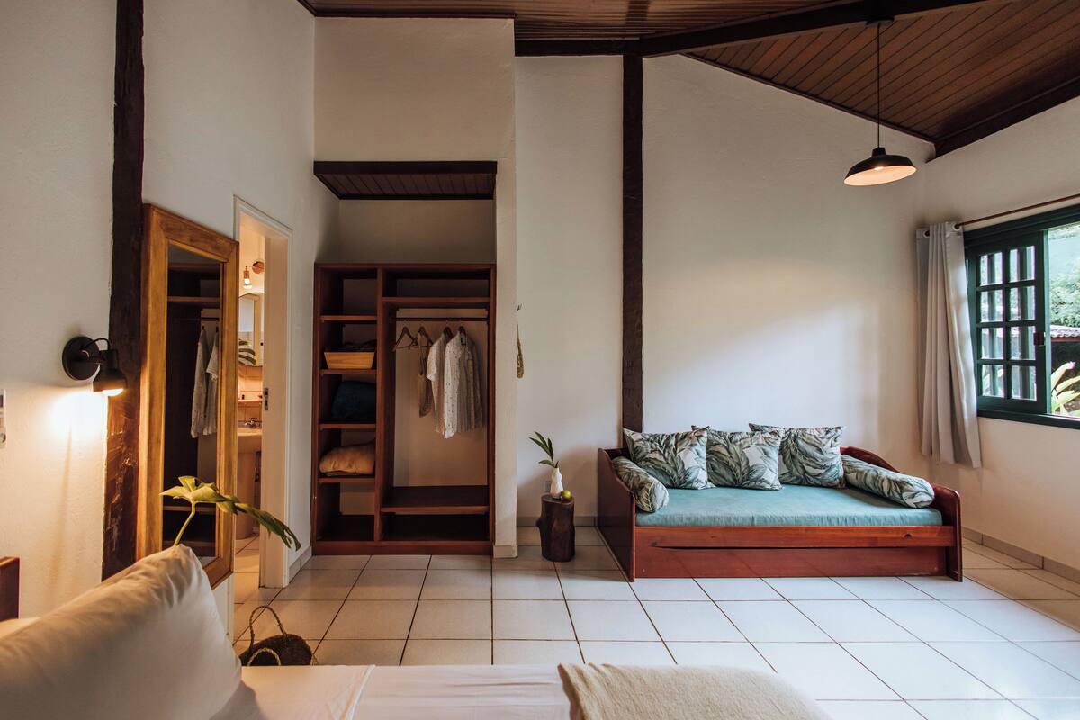 Una habitación con sofá de madera, una mesita, un espejo y un armario con ropa colgada.