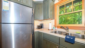 Una cocina con un refrigerador de acero inoxidable, una ventana que ofrece vistas al verdor, y un lavamanos con una toalla colgada del grifo.