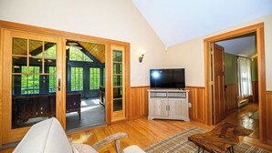 Un salón amplio con un televisor grande, piso de madera y vistas a una terraza exterior a través de puertas de vidrio.