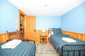 Ein Zimmer mit zwei Betten, einem Holzstuhl, einem kleinen Tisch mit einer Lampe und einer Tür zu einem weiteren Raum.