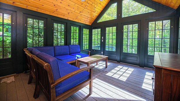 Una terraza de madera con un sofá azul y una mesa, ventanas amplias y un ventilador de techo.