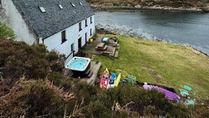 Un edificio blanco con techo inclinado cerca de una masa de agua, un área verde con jacuzzi y kayaks.