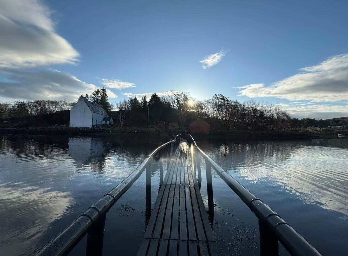 A wooden dock extending over calm water towards a white building with a sloped roof, surrounded by trees and a clear sky.