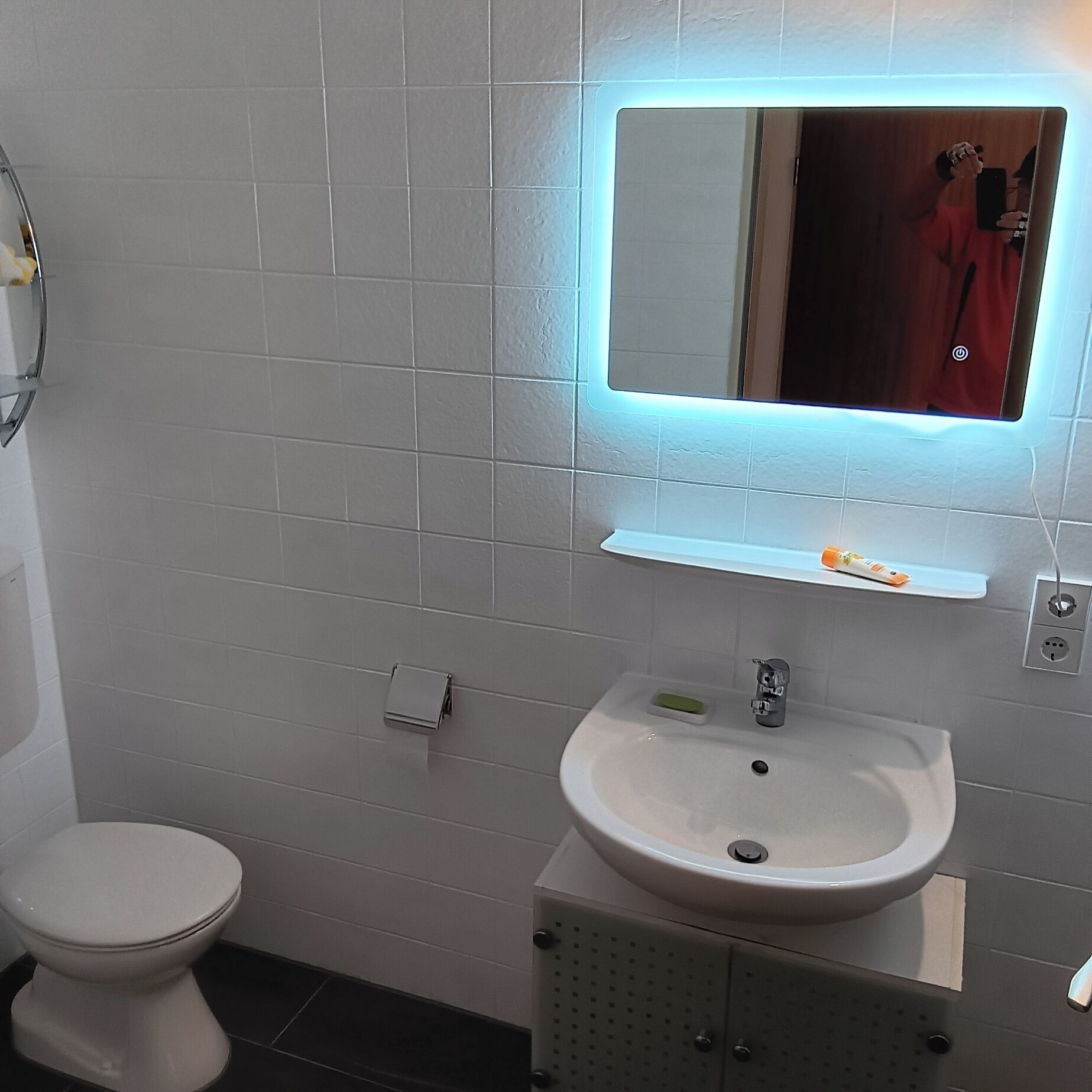 Un baño moderno con un inodoro blanco, un lavabo con espejo y una pequeña estantería con artículos de aseo.
