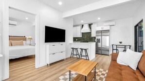 Un apartamento moderno con sala de estar, cocina y dormitorio. Incluye un sofá, un televisor, una mesa de madera y una isla de cocina con taburetes.