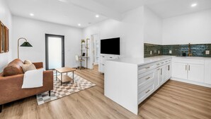 Una sala moderna con un sofá marrón, una mesa de centro y un televisor. Incluye una zona de cocina con armarios blancos y salpicadero de azulejos.