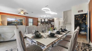 A modern dining area with a long table set for six, surrounded by comfortable seating. A television is mounted on the wall, and a staircase leads to an upper level. - Immersive Activities, Arcade, Theatre Sleeps 24 (Davenport)