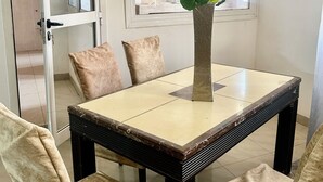 Uma sala de jantar com uma mesa quadrada, quatro cadeiras, um vaso de planta e uma janela com persianas.