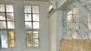 Quarto com cama, travesseiros, manta azul, parede com padrão marmorizado e janela com vista para palmeiras.