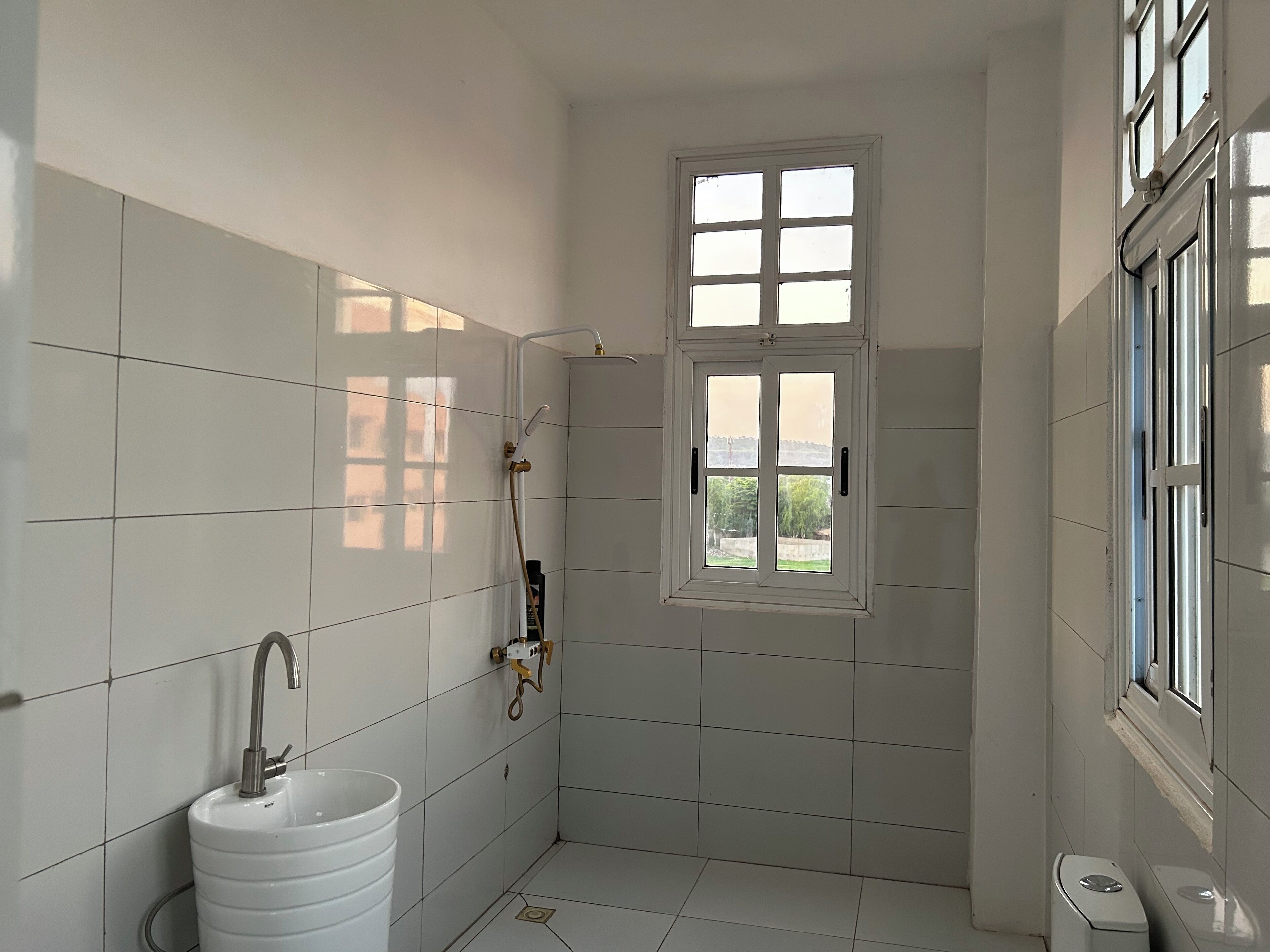Baño con azulejos blancos, lavamanos, ventana y ducha.
