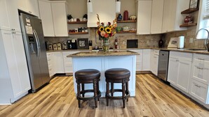 A modern kitchen with white cabinets, a stainless steel refrigerator, a granite countertop, and two wooden stools. - Heated Pool King Bed Jacuzzi Bath Arcade (Winston-Salem)